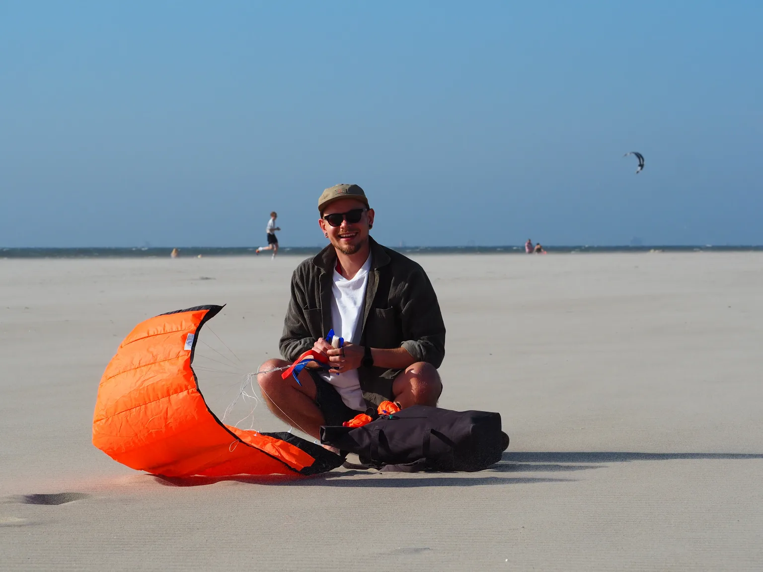 Mathias on a wide sandy beach by the sea, holding the handles of an orange stunt kite laid out on the sand